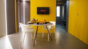 A modern dining area with a table set for two, a flat-screen TV mounted on the wall, and a yellow accent wall. - Fiveplace Design Suite & Apartments (Trapani)