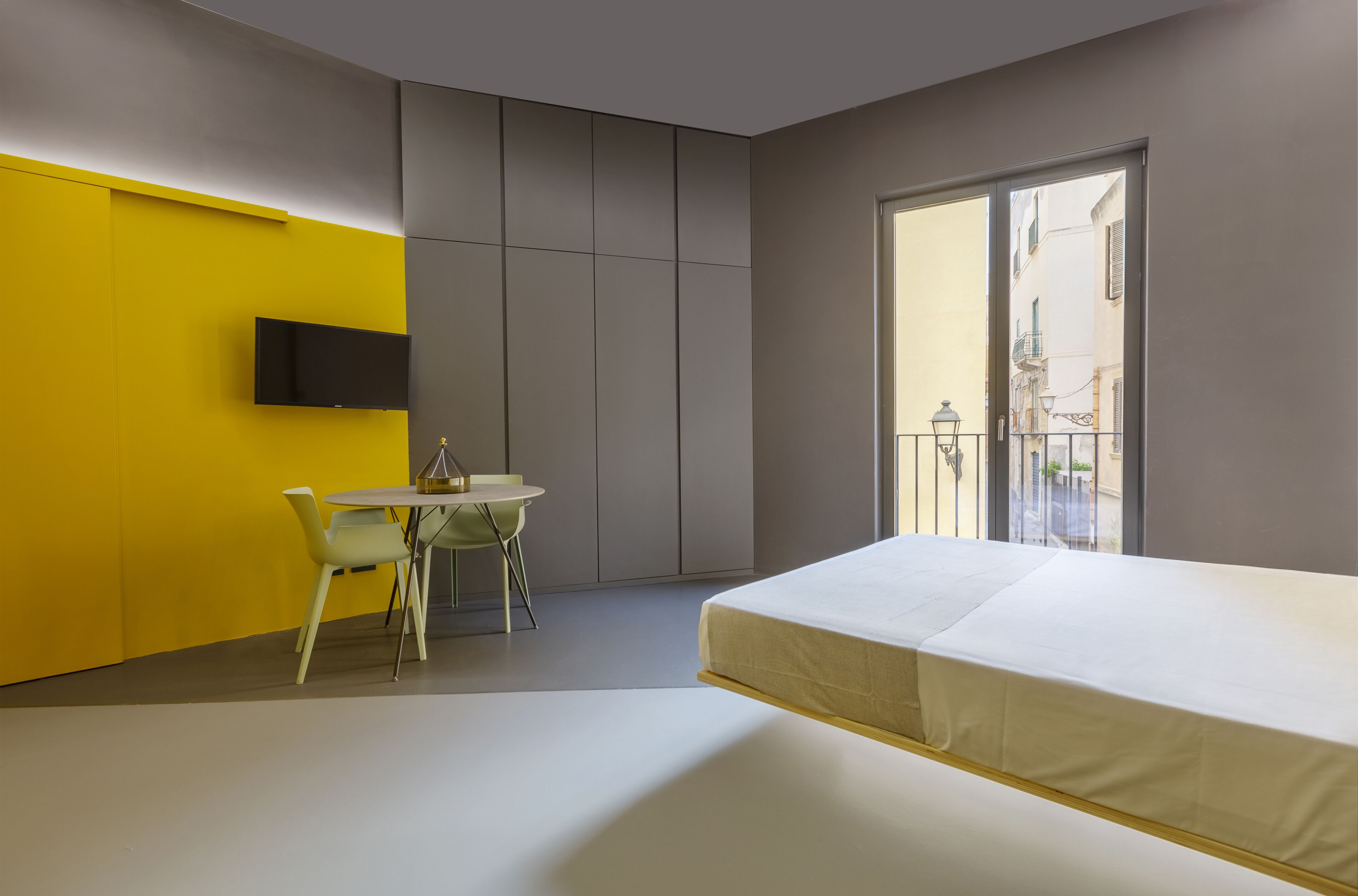 A modern bedroom with a bed, a small table with chairs, a TV mounted on a yellow wall, and a balcony with a view of buildings.