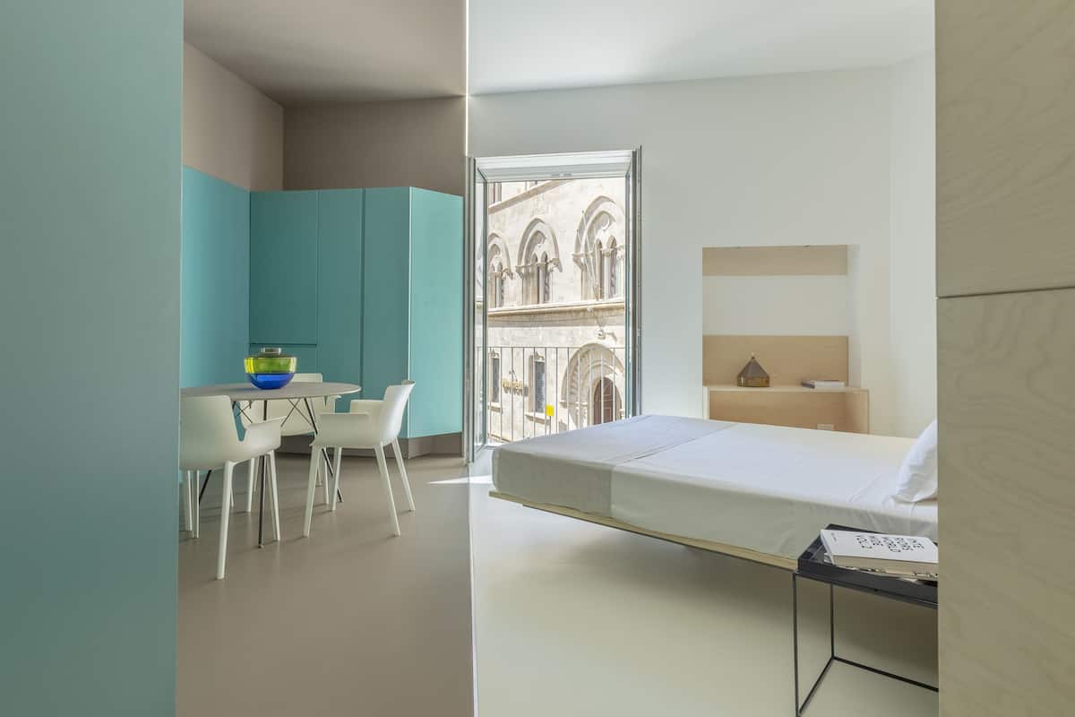 A modern hotel room with a bed, a desk with chairs, a small table with a bowl, and a view of a building through a glass door.