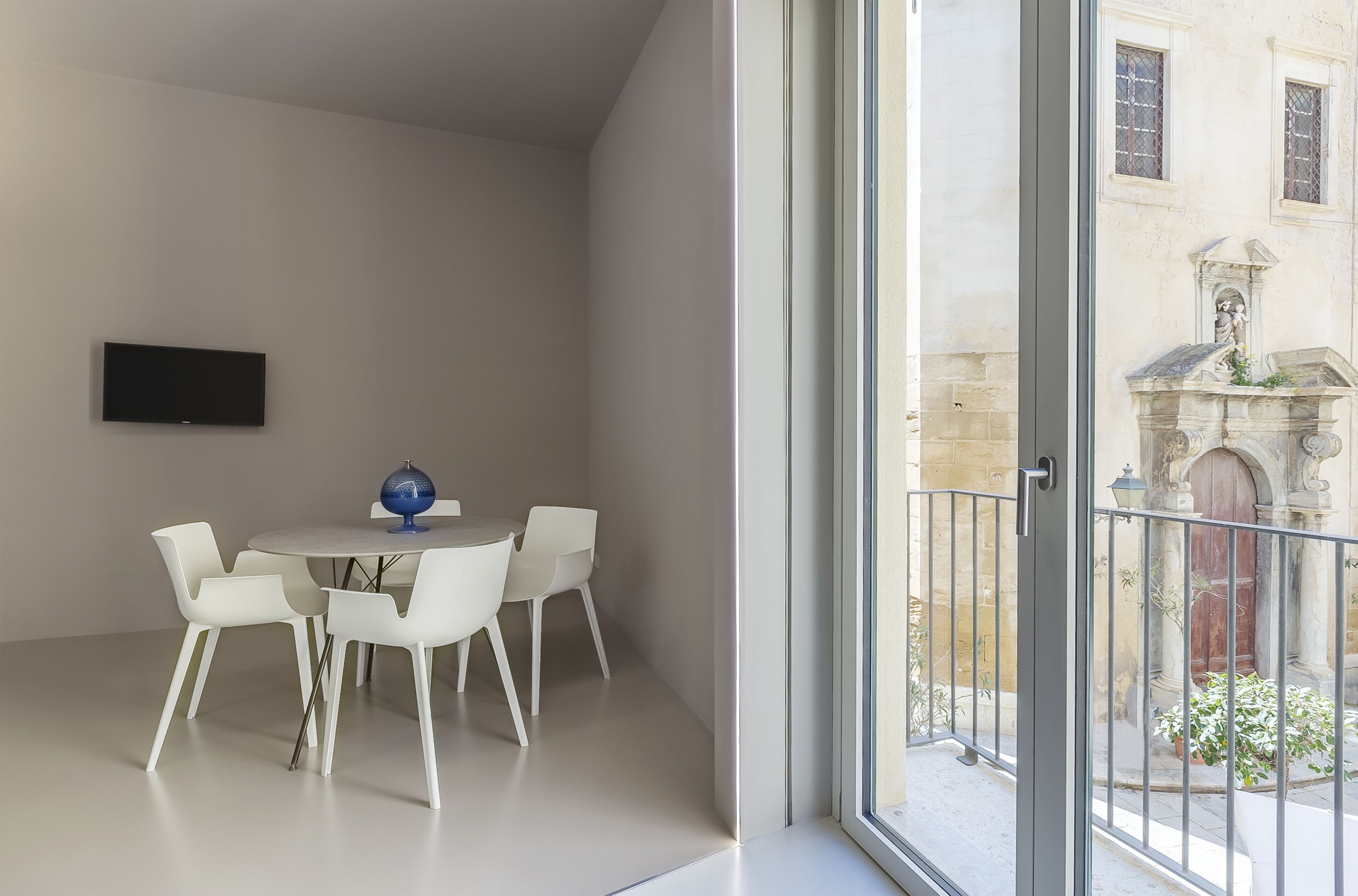 A modern room with a round dining table, white chairs, a flat-screen TV, and a large window overlooking a courtyard with a statue and plants.