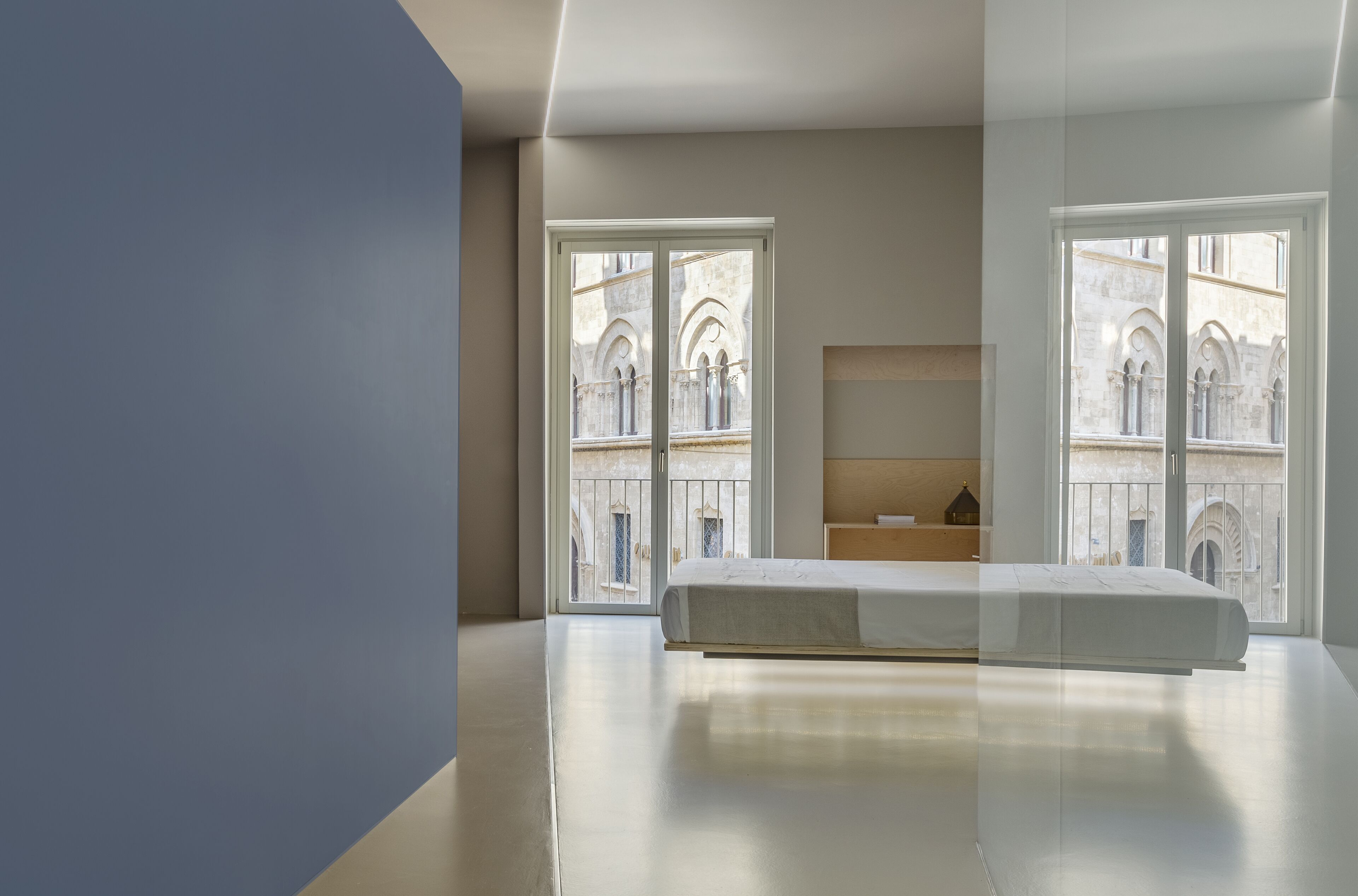 Una camera da letto moderna con un letto grande, un comodino in legno e vista su un edificio storico attraverso ampie finestre.