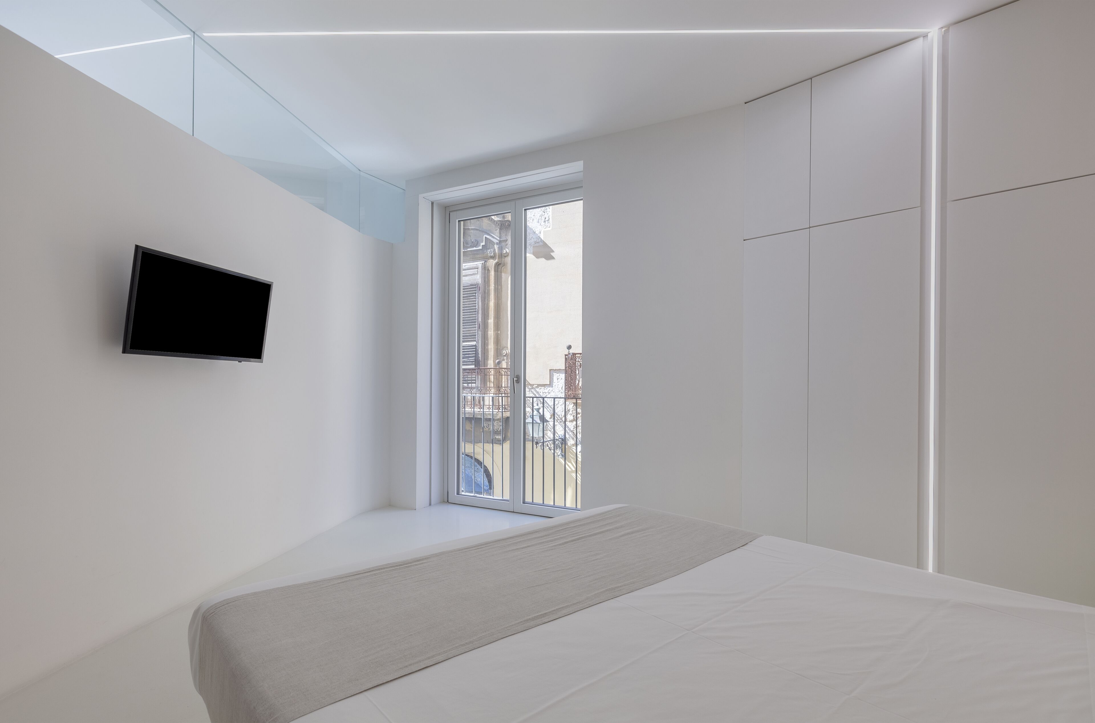 A modern bedroom with a bed, a flat-screen TV mounted on the wall, and a large window providing a view of the outside.