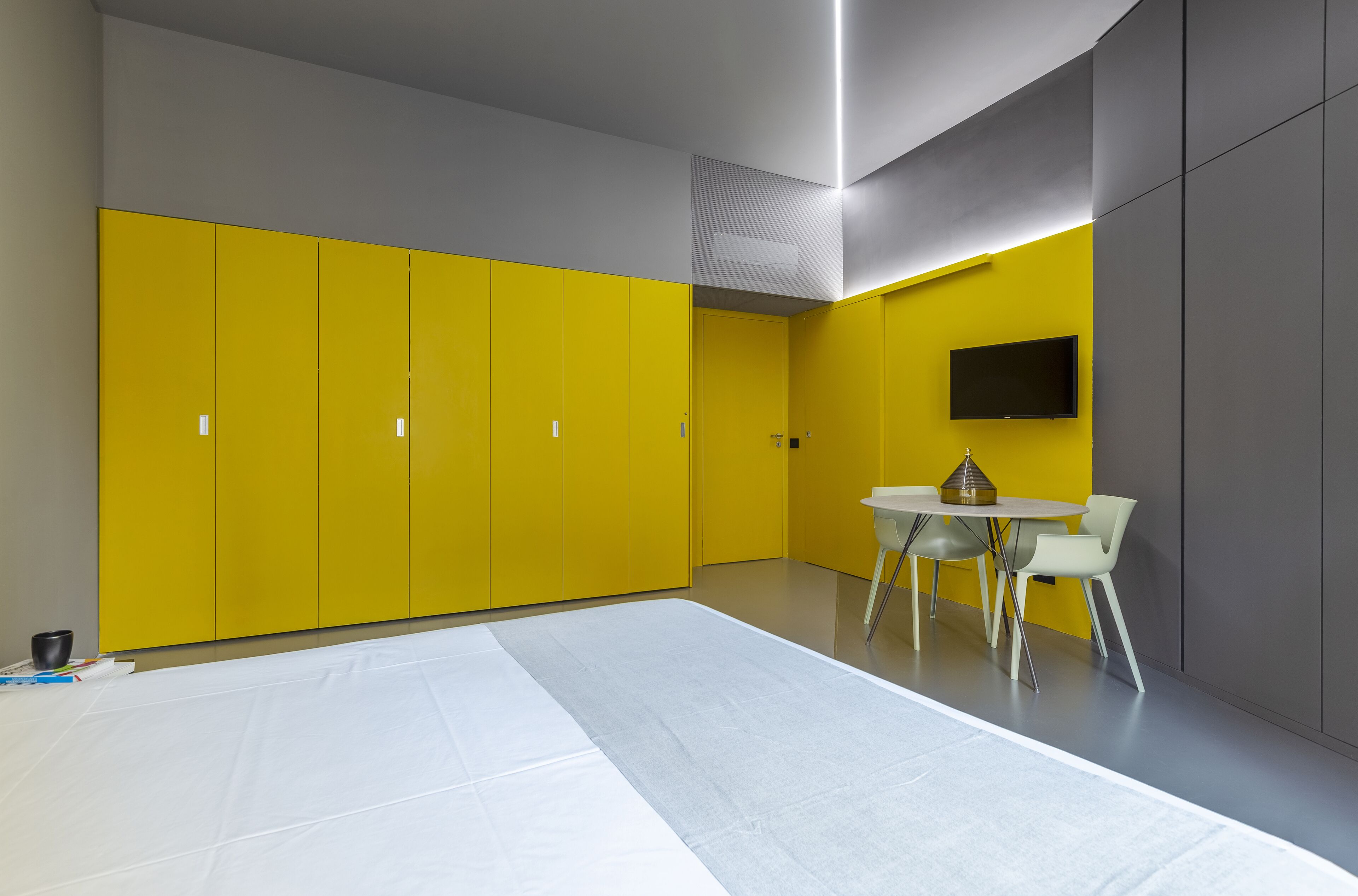 A modern room with yellow and grey walls, a white table with chairs, and a flat-screen TV mounted on the wall.