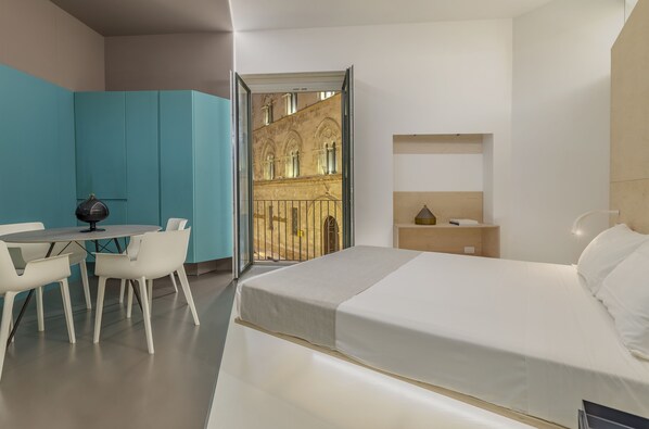 A modern hotel room with a bed, a desk with chairs, a small table, and a view of an outdoor courtyard. - Fiveplace Design Suite & Apartments (Trapani)