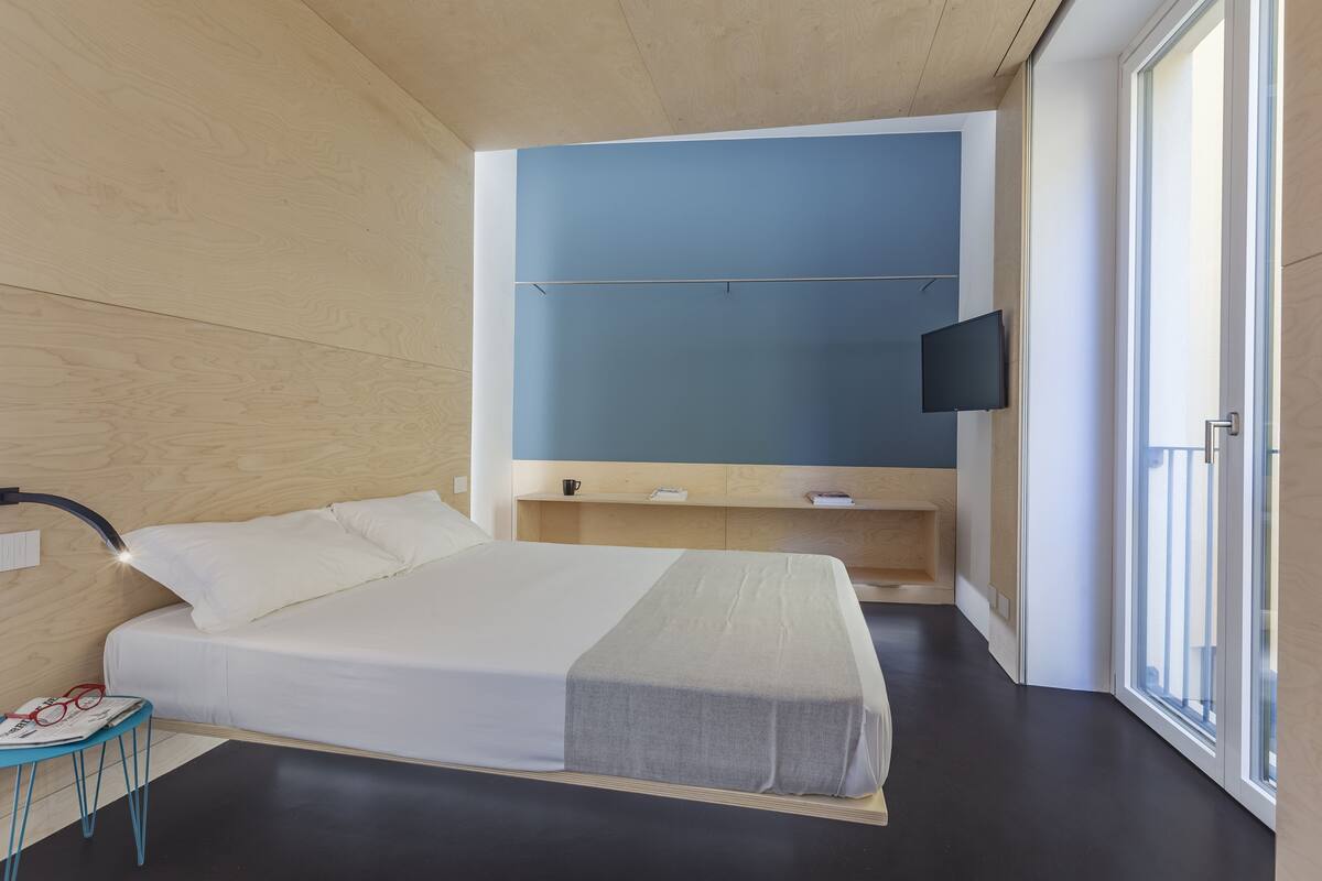A modern bedroom with a bed, a TV mounted on the wall, and a window with a view.