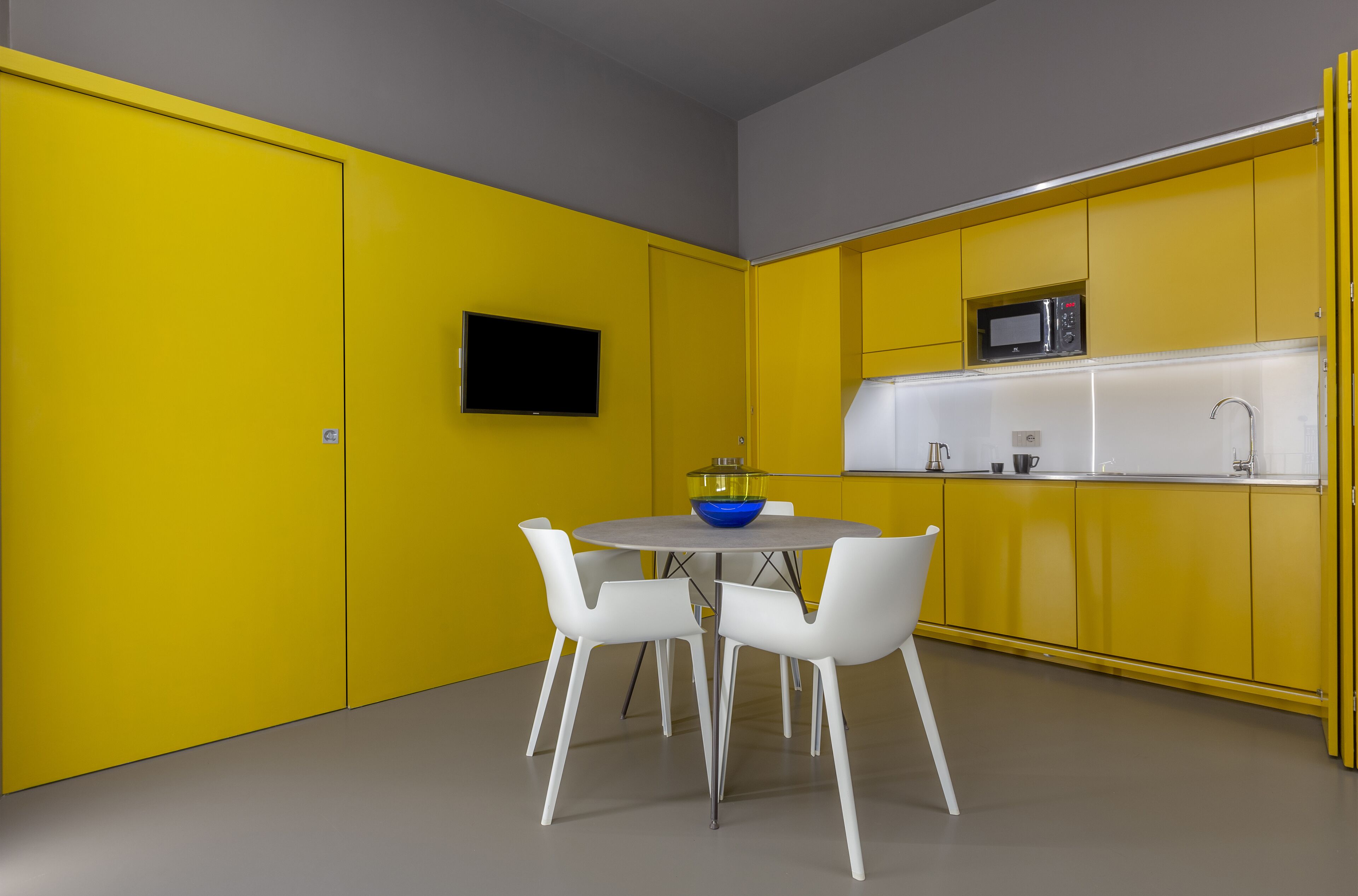 Una cocina moderna con armarios amarillos, encimera gris y sillas blancas alrededor de una mesa redonda con un cuenco azul.