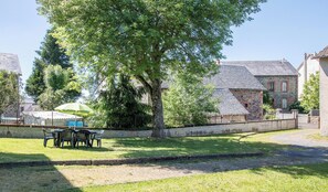 A stone building with a tiled roof, a tree in front, and a table with chairs under an umbrella. - Gîte des Trois Vallées (MESSEIX)