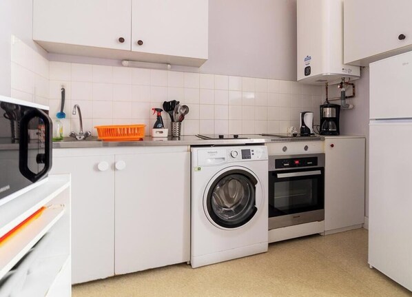 A compact kitchen with a washing machine, oven, and coffee maker. - Gîte des 3 Vallées - Messeix (MESSEIX)