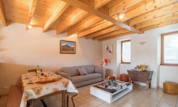 Un salon chaleureux avec un plafond en bois, un canapé, une table à manger recouverte d’une nappe à motifs floraux et une table basse agrémentée de livres et de fleurs.