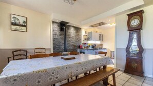 A dining area with a long table, chairs, a bench, a clock, and a kitchen in the background.