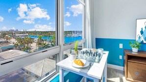 Ett rum med ett stort fönster som ger utsikt över en småbåtshamn och stadens silhuett. På ett bord står ett schackbräde, en croissant och en kopp.