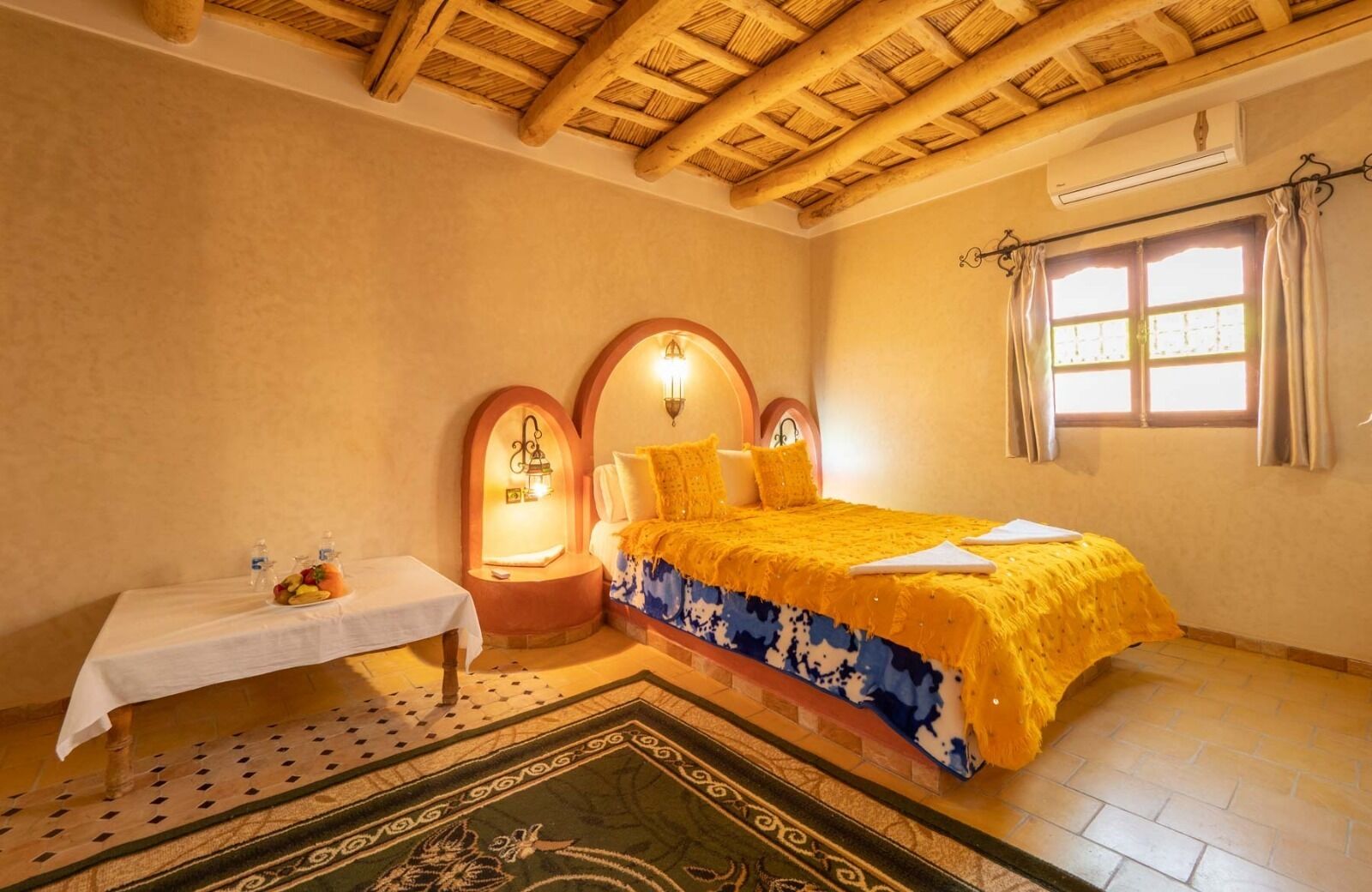 Un dormitorio con cama, una mesita de noche con frutero, una ventana con cortinas y un techo de madera.