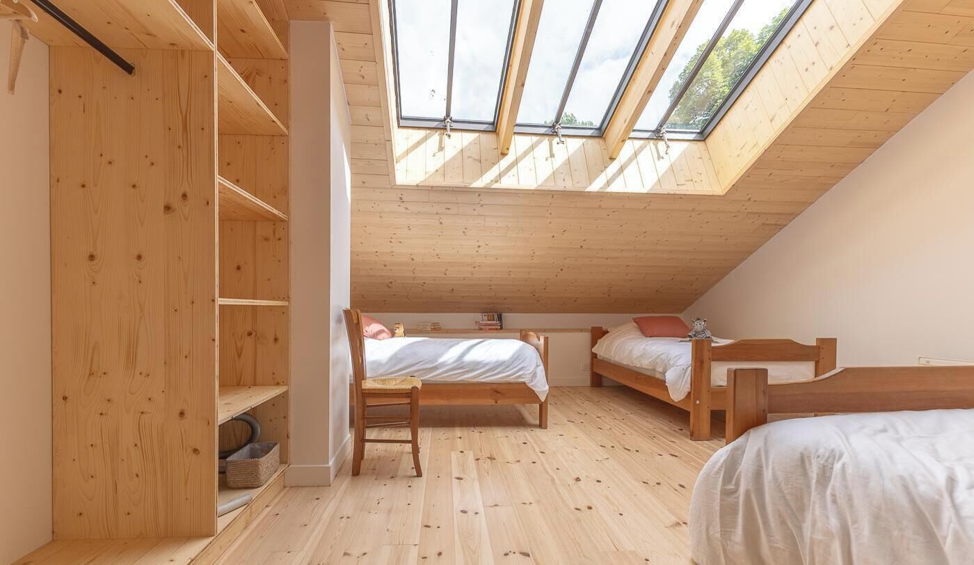 A room with a sloped ceiling, wooden flooring, two beds, a chair, and a skylight.