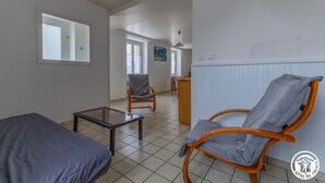 Quarto com cama, cadeira, mesa e radiador. Há uma janela e uma porta que dá para outro cômodo.