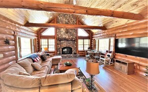 A log cabin interior with a stone fireplace, a large sofa, a television, and a dining area.