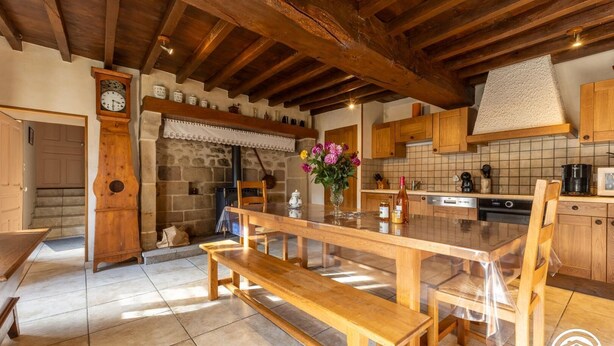 Una cocina rústica con vigas de madera, pared de piedra, mesa de madera con bancos, un reloj y una estufa.