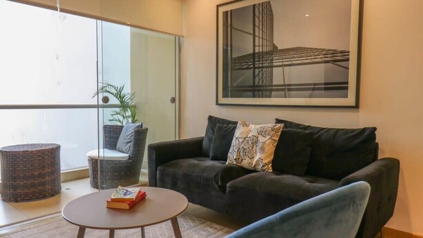 A modern living room with a dark sofa, a round coffee table, and a wicker chair. A large framed picture hangs on the wall.