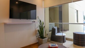 A modern living room with a flat-screen TV mounted on the wall, a potted plant, wicker furniture, and a view through large glass doors to an outdoor area.