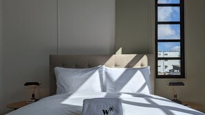 Una cama bien tendida con sábanas blancas, una toalla gris con un logotipo y una ventana con vistas a edificios.