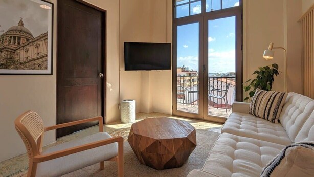 Un salón con sofá, una mesa de centro de madera, un televisor montado en la pared y un ventanal con vistas a edificios.
