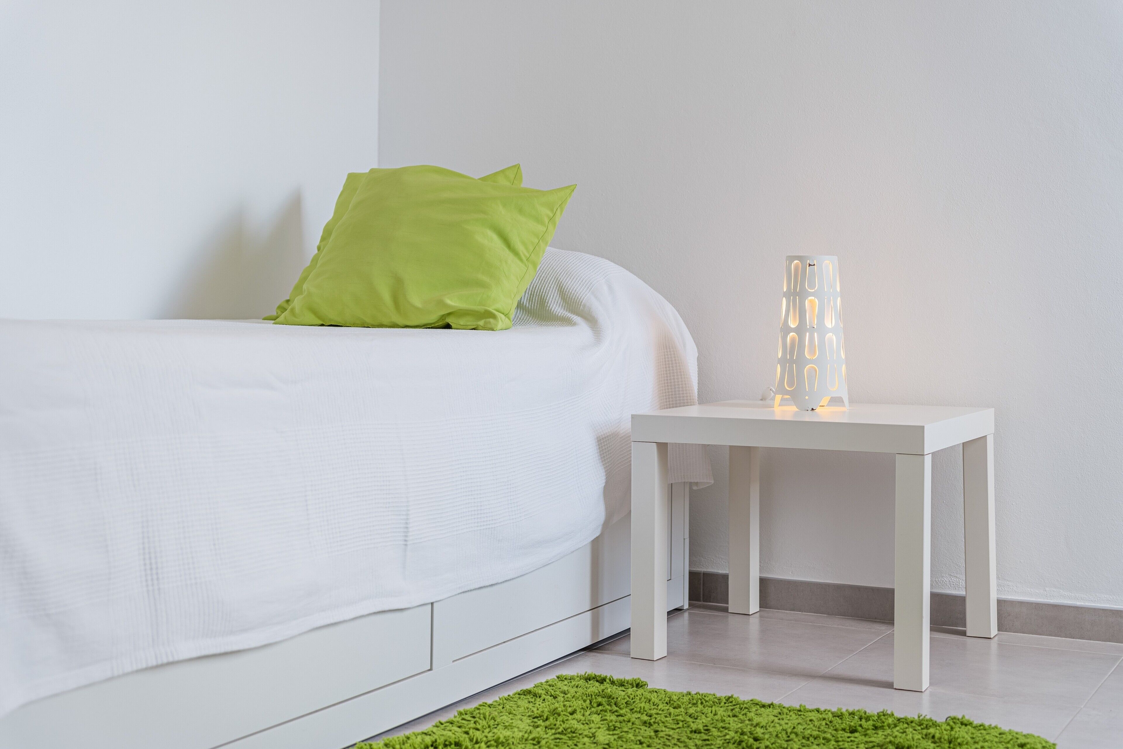 Un lit bien fait, avec un matelas blanc et des oreillers verts, une table de chevet blanche sur laquelle se trouve une lampe blanche, et un tapis vert sur le sol.