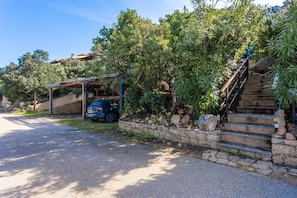A residential area with a stone staircase leading to a wooden deck, surrounded by lush greenery and a parked car. - Holiday Home "Villetta Duplex Breathtaking View" with Sea View, 2 Private Terraces and Wi-Fi (Costa Paradiso)