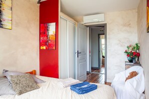 A bedroom with a bed, pillows, a red accent wall, a painting, and a hallway leading to another room.