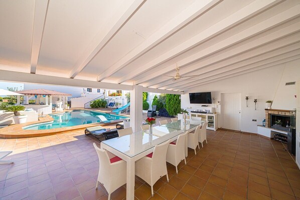 Un espacio exterior techado con piscina, una mesa de comedor con sillas, un televisor y una chimenea.