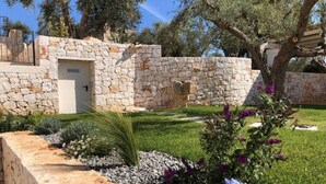 A stone building with a door and a window, surrounded by a well-maintained garden with grass, gravel, and flowering plants. - Trullo Mamamè (Puglia)