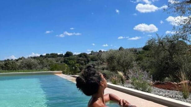 A person relaxing in a clear pool with a view of a natural landscape and distant buildings. - Trullo Mamamè (Puglia)