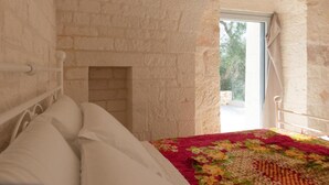A bedroom with a stone wall, a bed with white bedding, and a window with a view of trees. - Trullo Mamamè (Puglia)