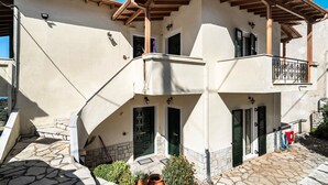Un edificio de dos pisos con entrada empaveda de piedra, un balcón con barandilla de hierro forjado y una puerta con contraventanas verdes.