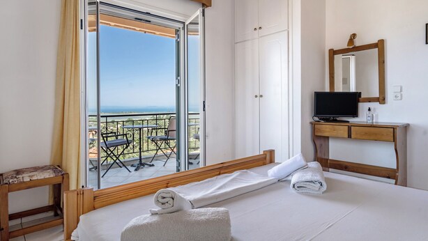 A hotel room with a bed, a television, a wooden desk, a mirror, and a balcony with a table and chairs. - Apartment "Lefkothea 4" with Sea View, Balcony & Wi-Fi (Lefkada)
