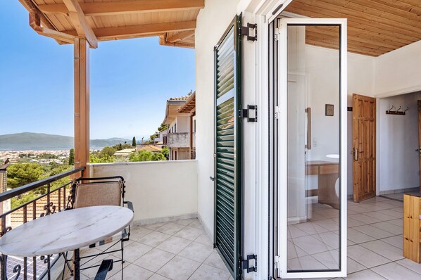 A balcony with a table and chairs, a view of the sea and buildings, and a wooden door leading inside.