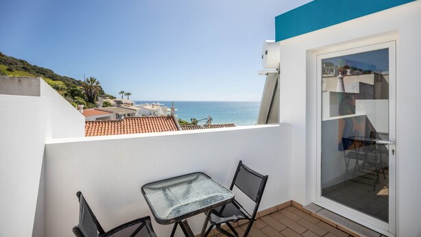 A balcony with a table and chairs, a glass door, and a view of the sea and buildings. - Holiday Home "Casa Da Paz" with Sea View, Private Terraces & Wi-Fi (Salema)