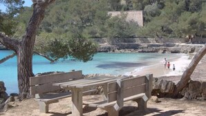 Una playa con un banco de madera, agua turquesa cristalina y un muro de piedra al fondo.