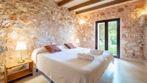 Un dormitorio con pared de piedra, vigas de madera, una cama con ropa de cama blanca, una mesita de noche con lámpara y vistas a través de un ventanal.