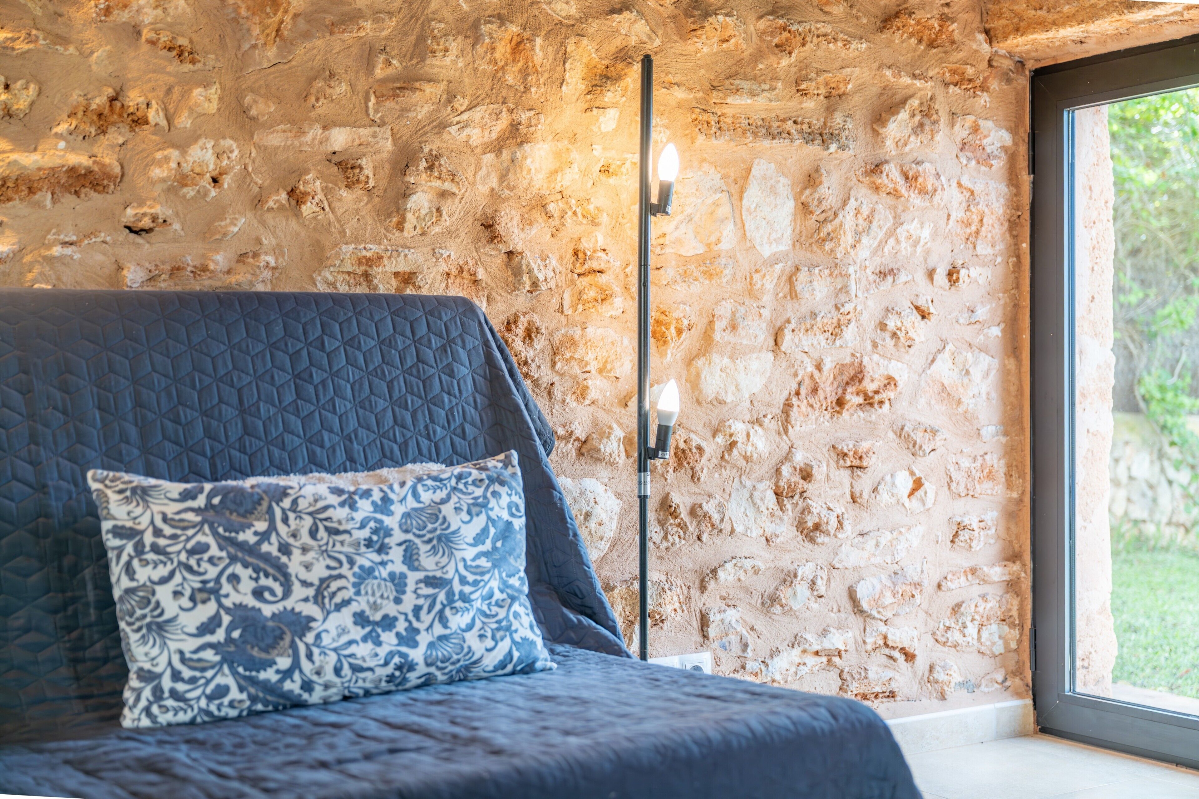 A room with a stone wall, a bed with a quilted cover, a patterned pillow, and a floor lamp.