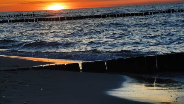 En strand med solnedgang over havet, bølger der slår ind mod kysten og en bølgebryder i det fjerne.