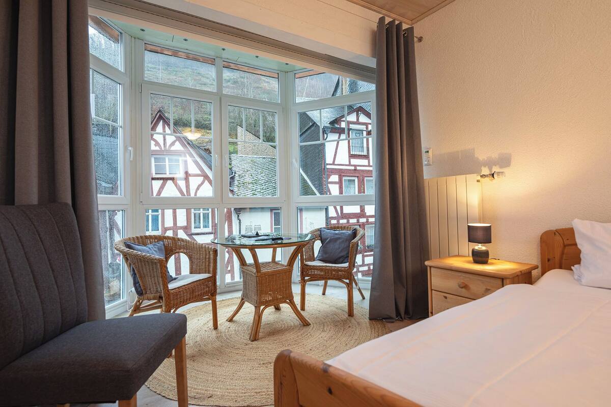 A hotel room with a large window, wicker furniture, a bed, and a nightstand with a lamp.