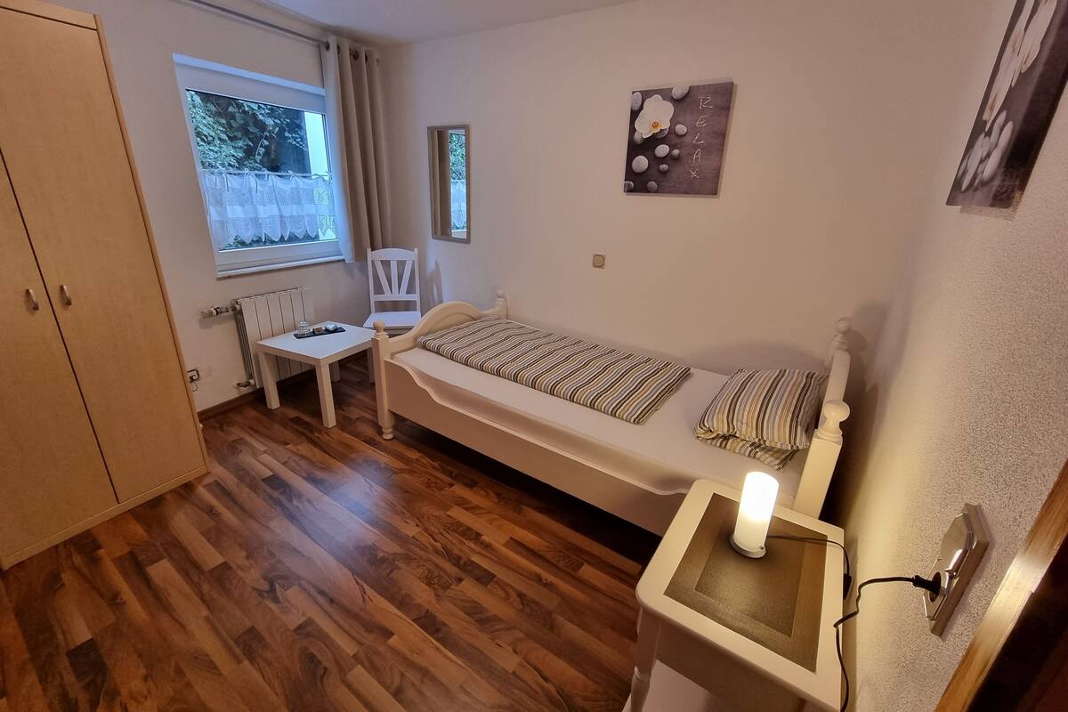 A small, tidy room with a wooden floor, a single bed with striped bedding, a small table, a chair, and a window with curtains.