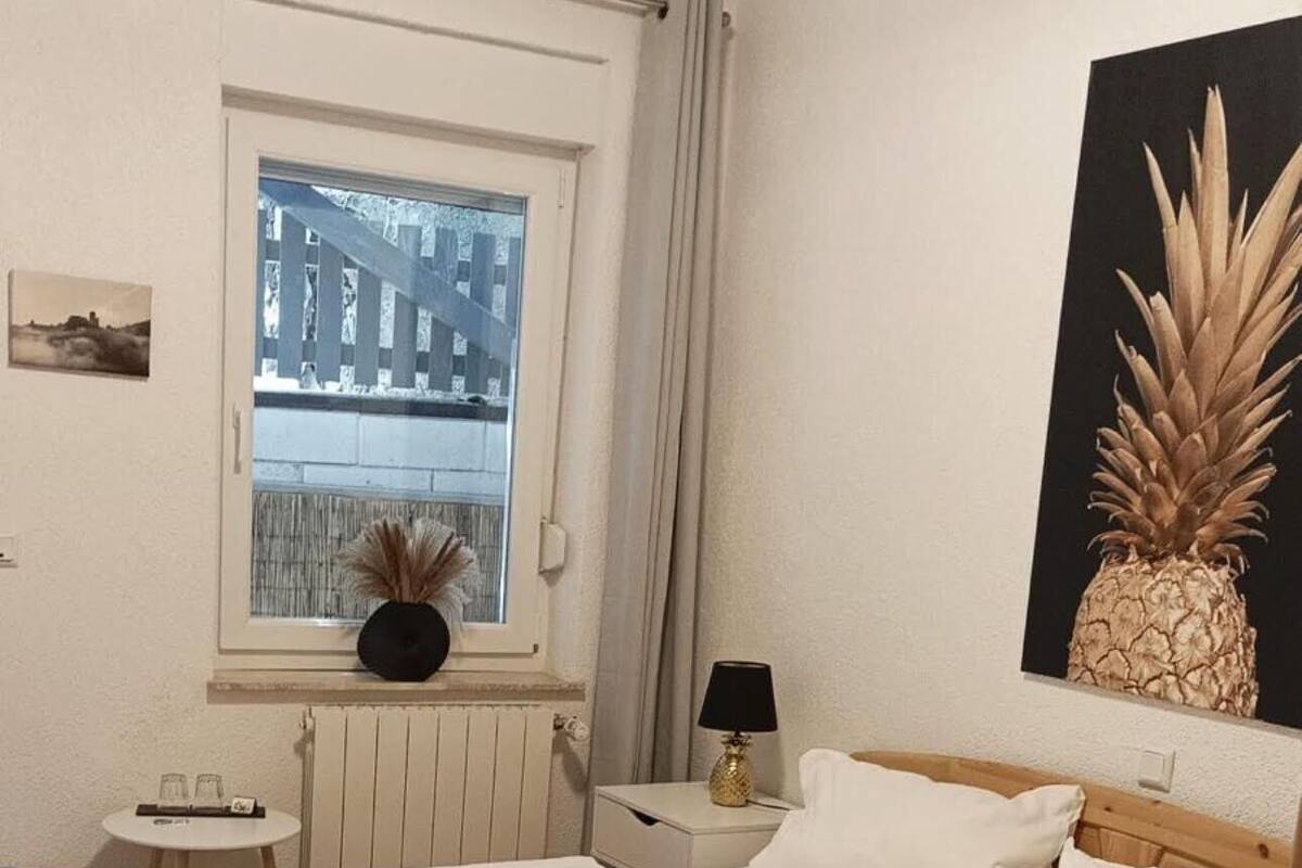 A bedroom with a wooden bed, a window with a view of a balcony, a small bedside table with a lamp, and a wall-mounted artwork of a pineapple.