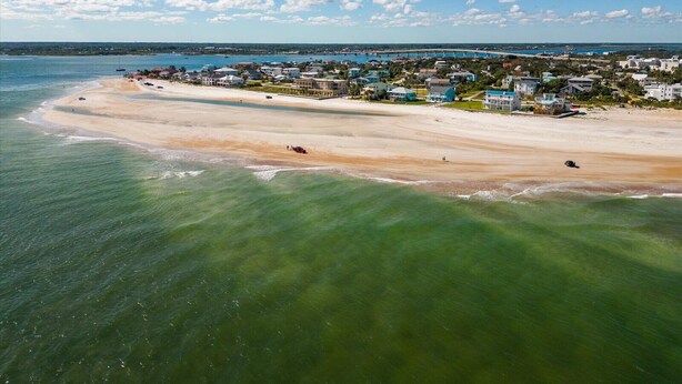 An aerial view of a coastal area with a sandy beach, a row of houses, and a clear blue sky.