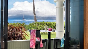 A patio with a table set for two, a view of the ocean, and a sailboat in the distance. - Beautiful ground floor unit right on the beach! (Lahaina)