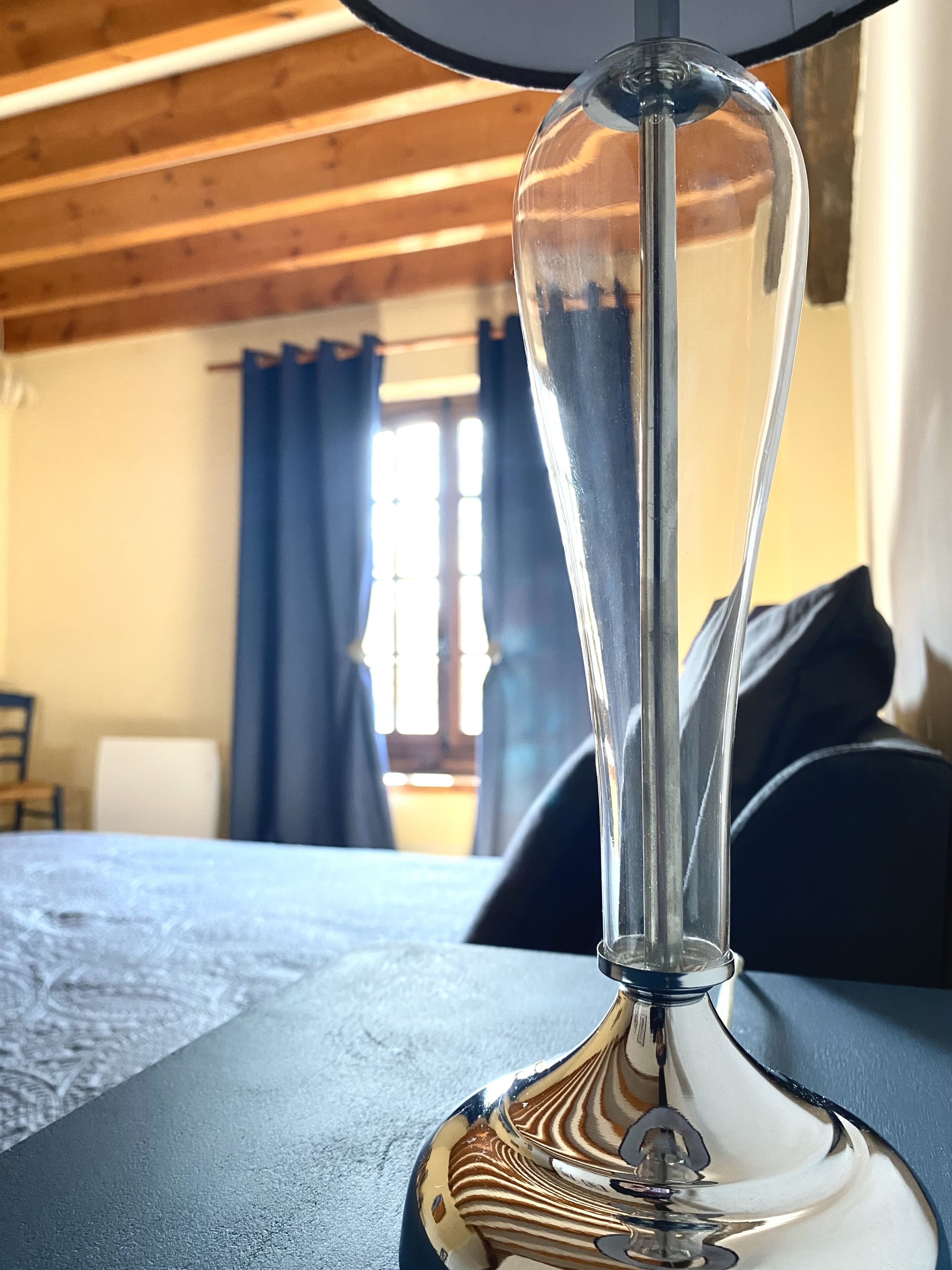 A clear glass table lamp with a metallic base, positioned on a table in a room with wooden beams on the ceiling and a window with curtains.