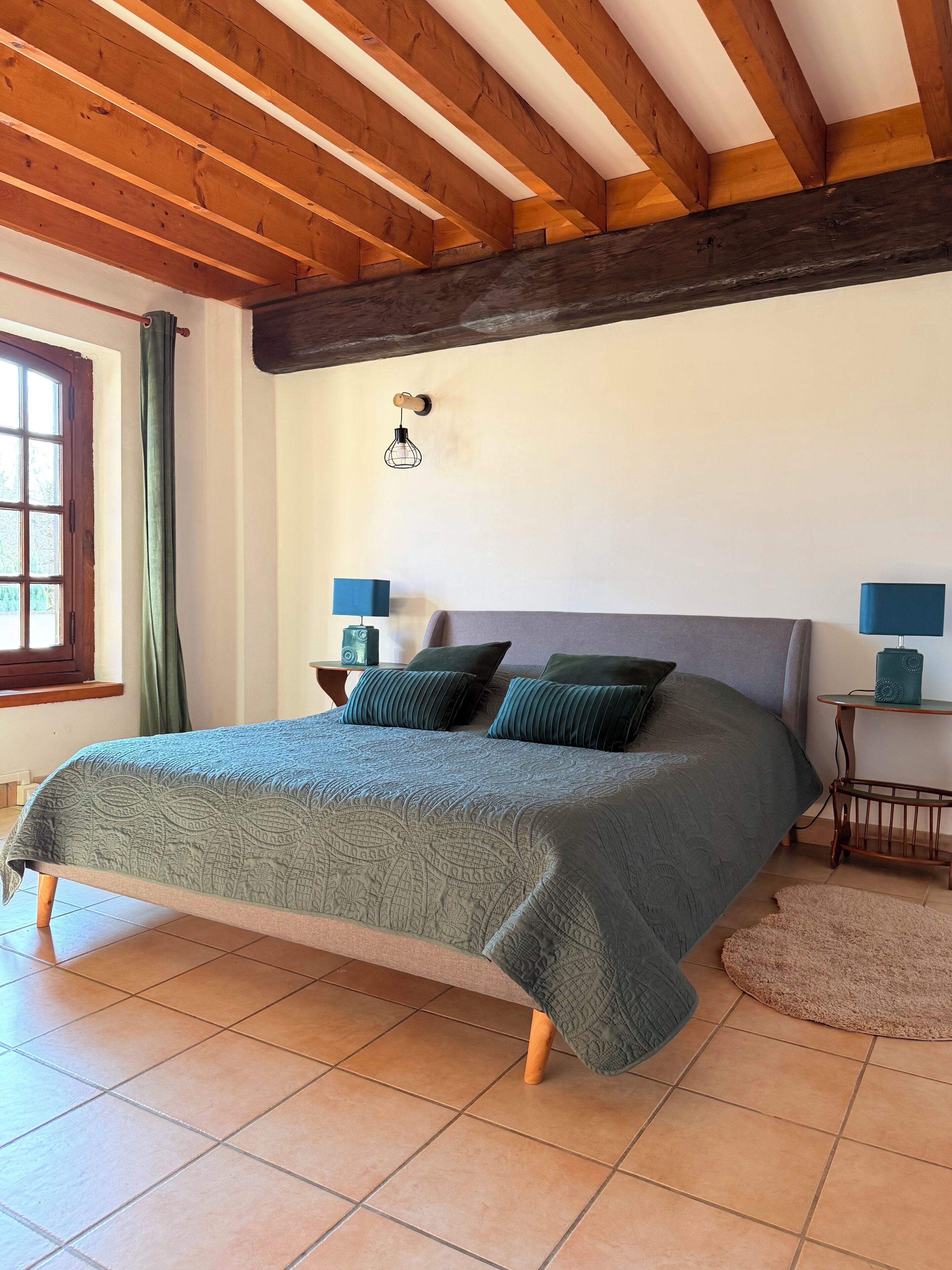Un dormitorio con techo de madera, una cama con colcha verde, dos mesitas de noche con lámparas y una ventana con cortinas.