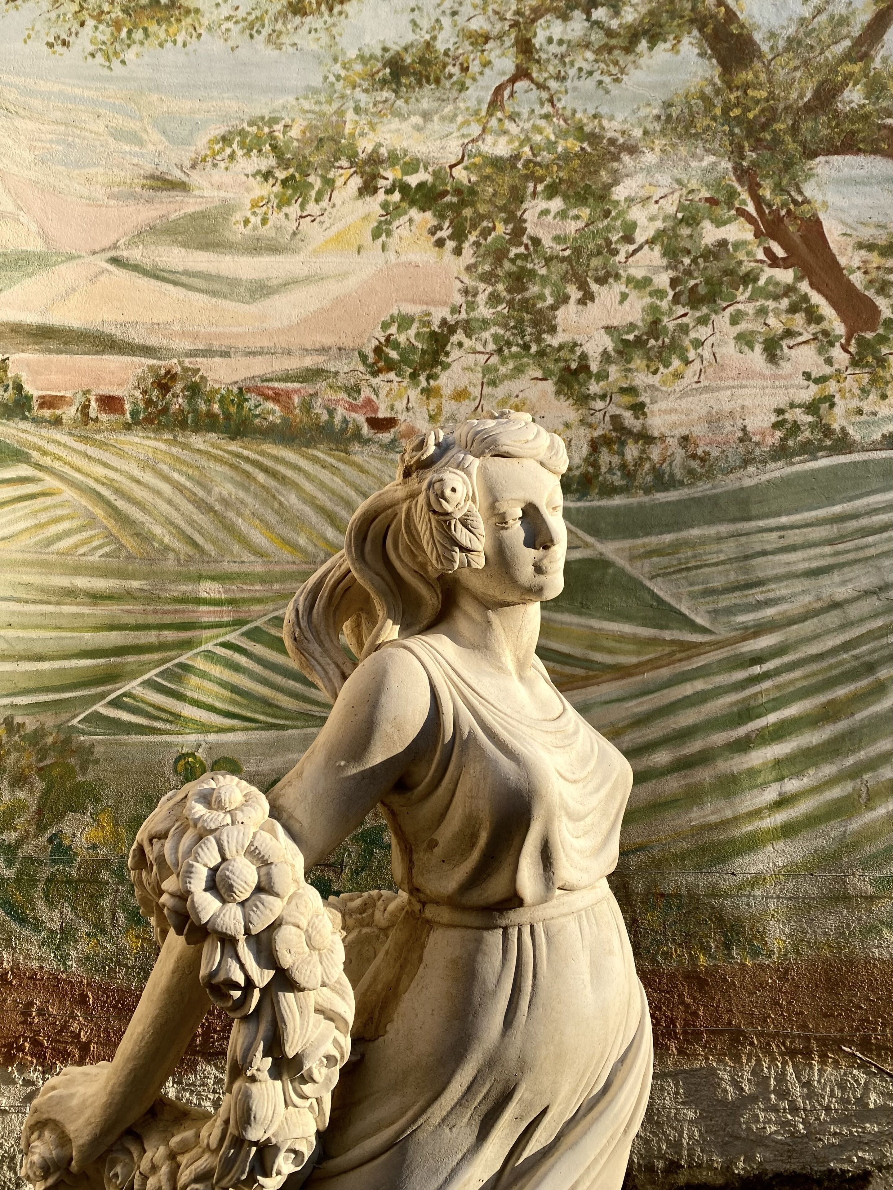 Una estatua de una mujer sosteniendo un racimo de uvas, con colinas ondulantes y un árbol de fondo.