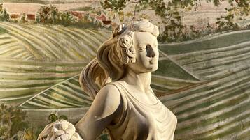 Una estatua de una mujer sosteniendo un racimo de uvas, con colinas ondulantes y un árbol de fondo.