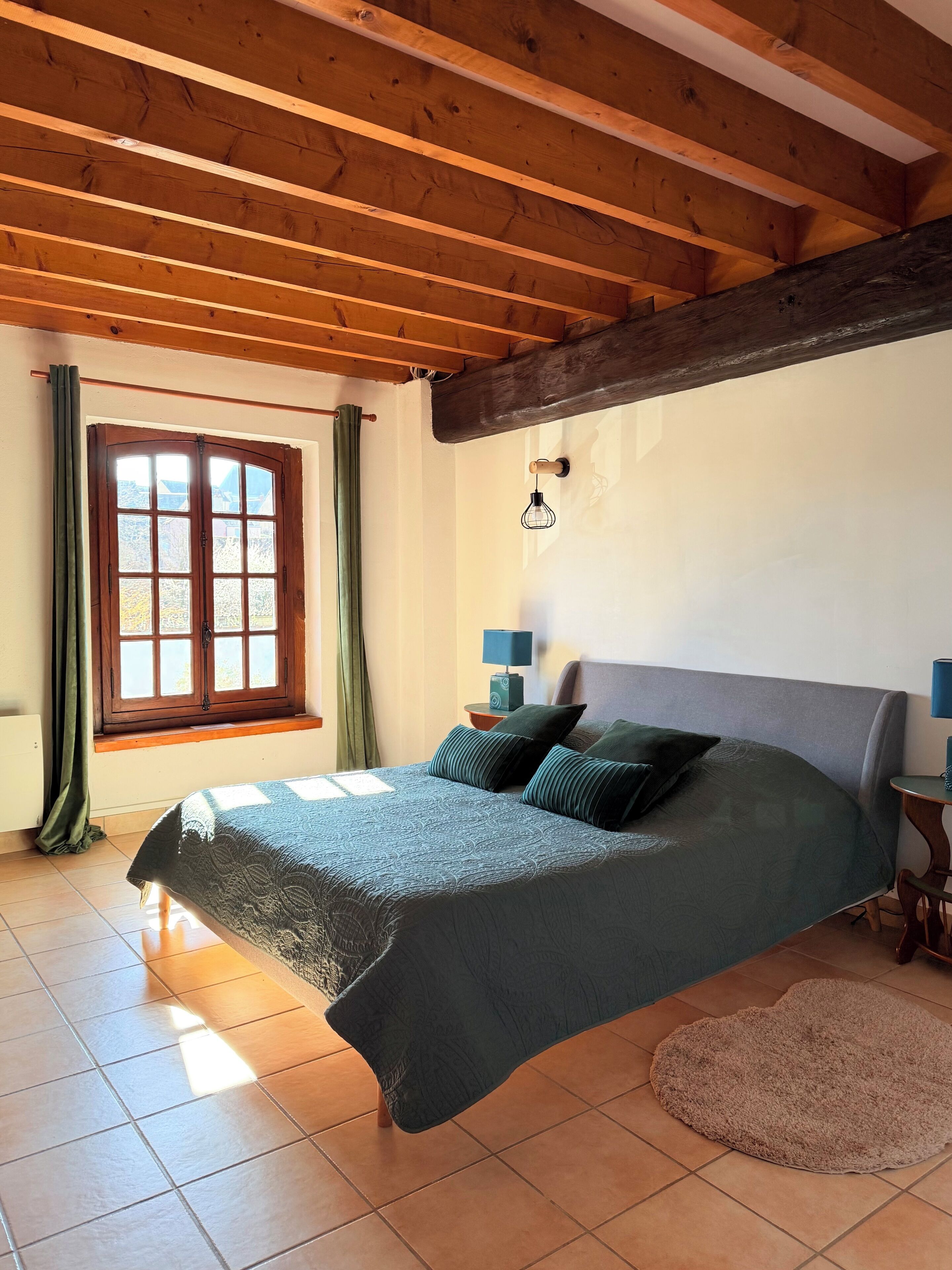 A bedroom with a wooden ceiling, a large bed with a green bedspread, two bedside tables with lamps, a window with curtains, and a view of the outside.