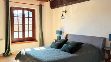A bedroom with a wooden ceiling, a large bed with a green bedspread, two bedside tables with lamps, a window with curtains, and a view of the outside.